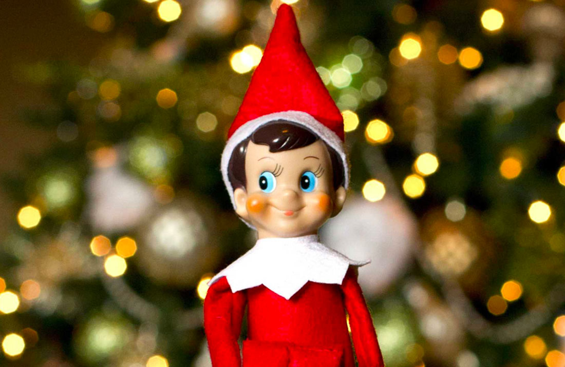The Elf on the Shelf smiling and looking cheeky, with the lights of a Christmas tree blurred in the background.
